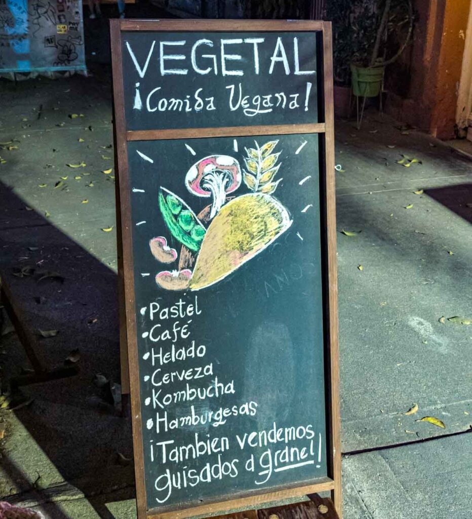 The outside menu board at Vegetal, a Mexico City vegan restaurant features a hand drawing of a vegan taco with a mushroom, peas, beans, and a plant coming out of the taco. Below is a list of the offerings including vegan cake and vegan ice cream.