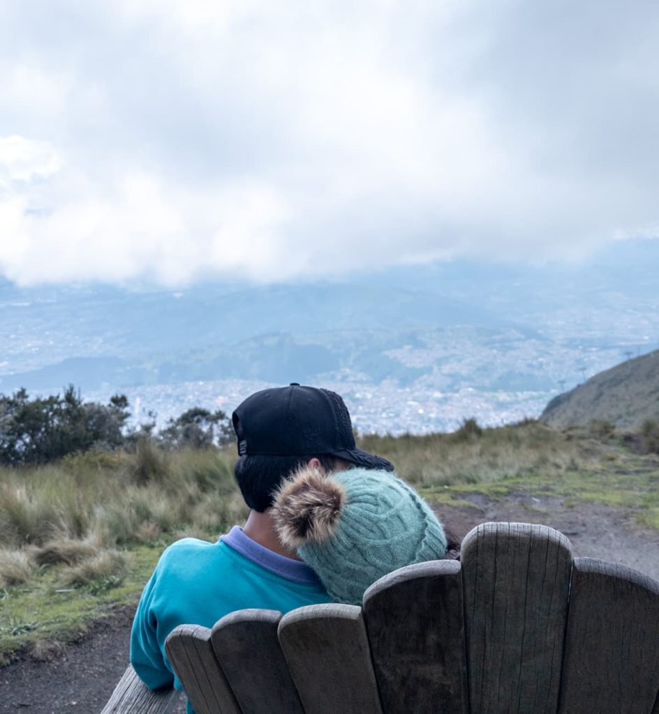 A couple sits closely on a wooden bench overlooking the city of Quito from a high-altitude viewpoint near the top station of the Teleférico.