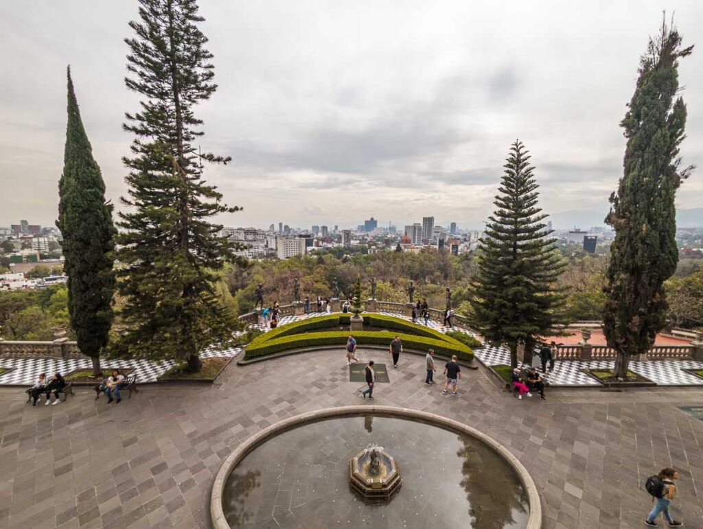 Views of Mexico City from Chapultepec Castle in the background as people wander on the terrace in front of a large circular fountain.