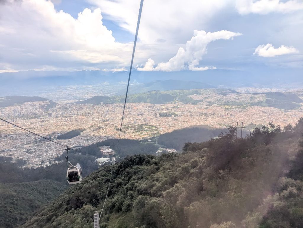 Looking out of the cable car in Quito as it descends over lush green hills with a panoramic view of the city sprawling below and dramatic clouds above.