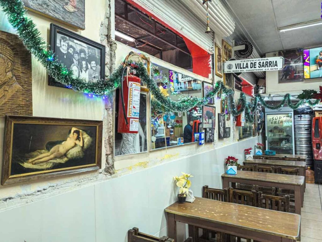Various images and decor hang on the walls at Villa de Sarria bar in Mexico City. Along the wall are skinny rectangular wooden tables and chairs.