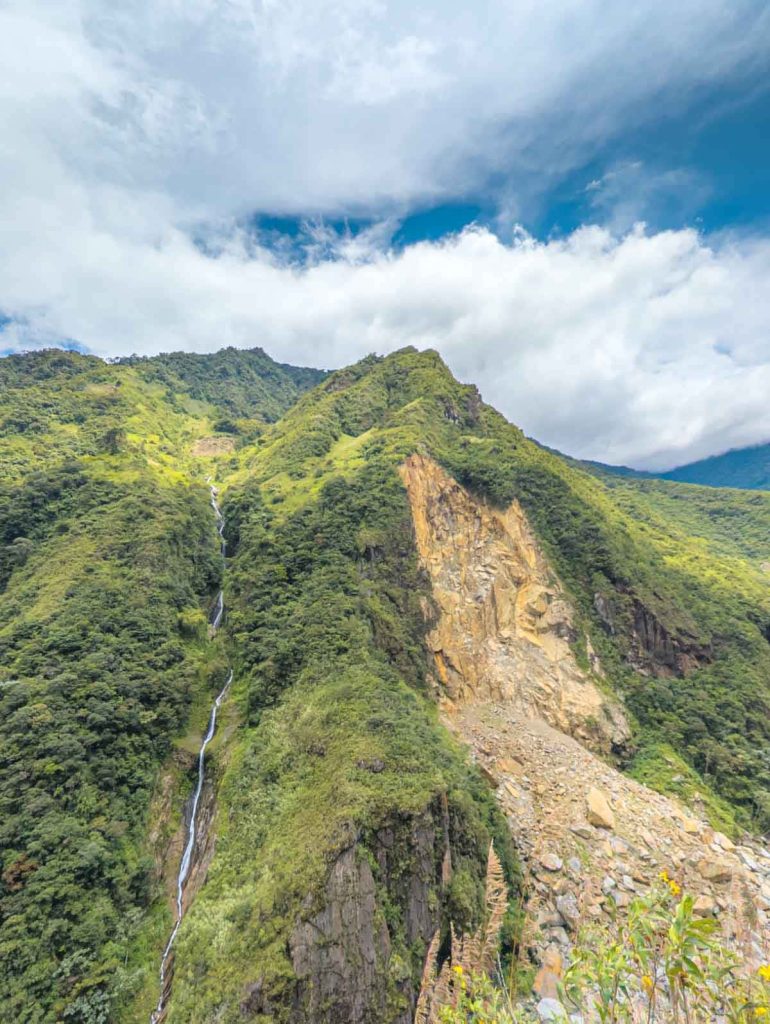 A thin waterfall flowing down a steep, bright green mountainside, surrounded by jagged rock formations along the Ruta de las Cascadas in Baños.