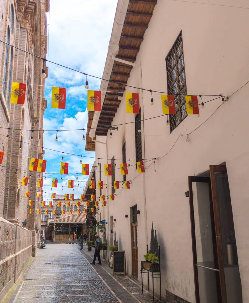 A narrow cobblestone street in Cuenca Ecuador lined with colonial buildings and strung with small red and yellow flags, located just behind the New Cathedral.