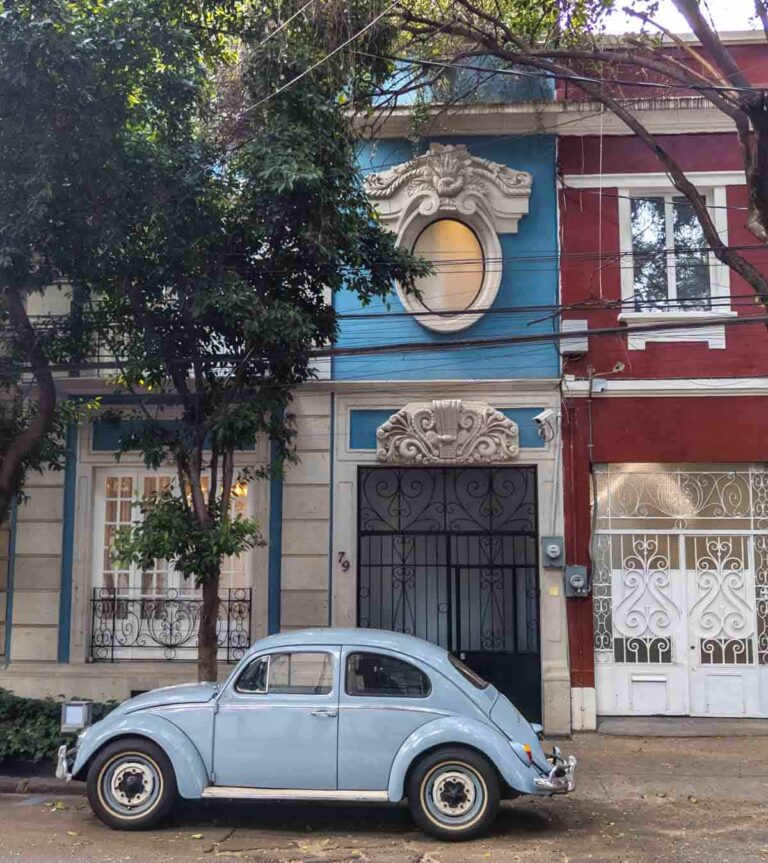 A light blue VW bug parked on a street in front of buildings featuring blue and red with ornate details.