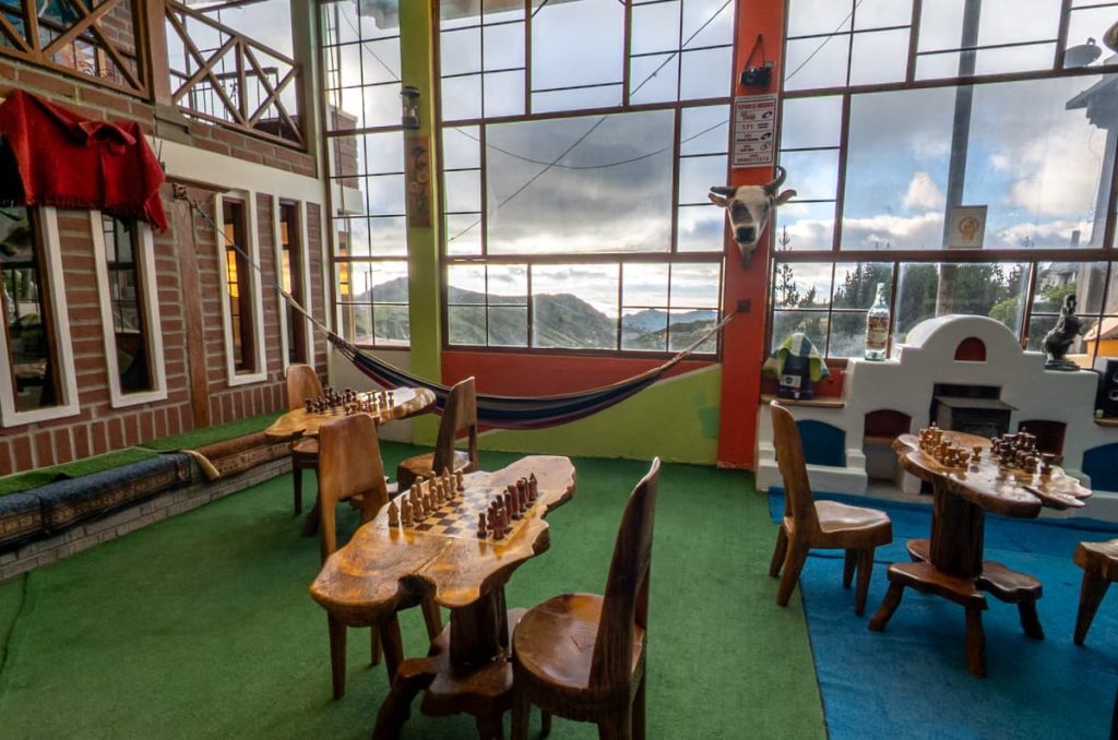A cozy indoor space at one of the best places to stay in Quilotoa Ecuador features handcrafted wooden chess tables, a hammock, and panoramic mountain views through large windows.