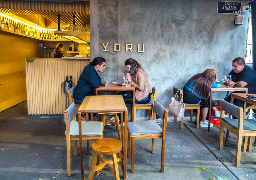 Couples sit at wooden tables outside in front of Yoru, which serves some of the best sushi in Mexico City. The modern building is made of concrete with gold lettering.