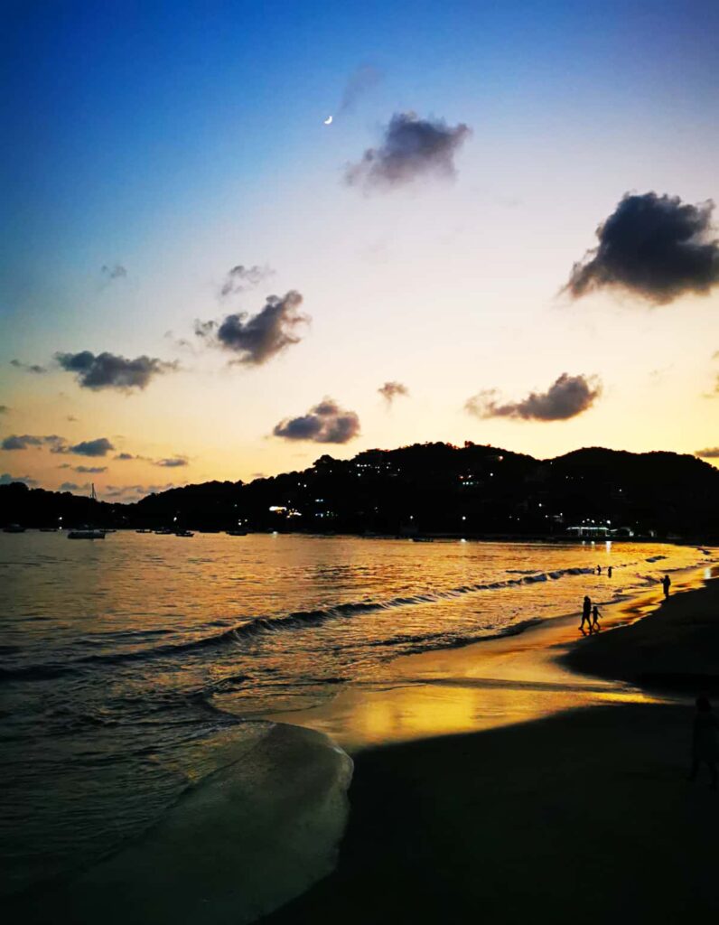 A crescent moon emerges in the sky as the golden hues of the end of sunset cast over the beach in Zihuatanejo. Several people continue to play in the waves and walk along the shore.