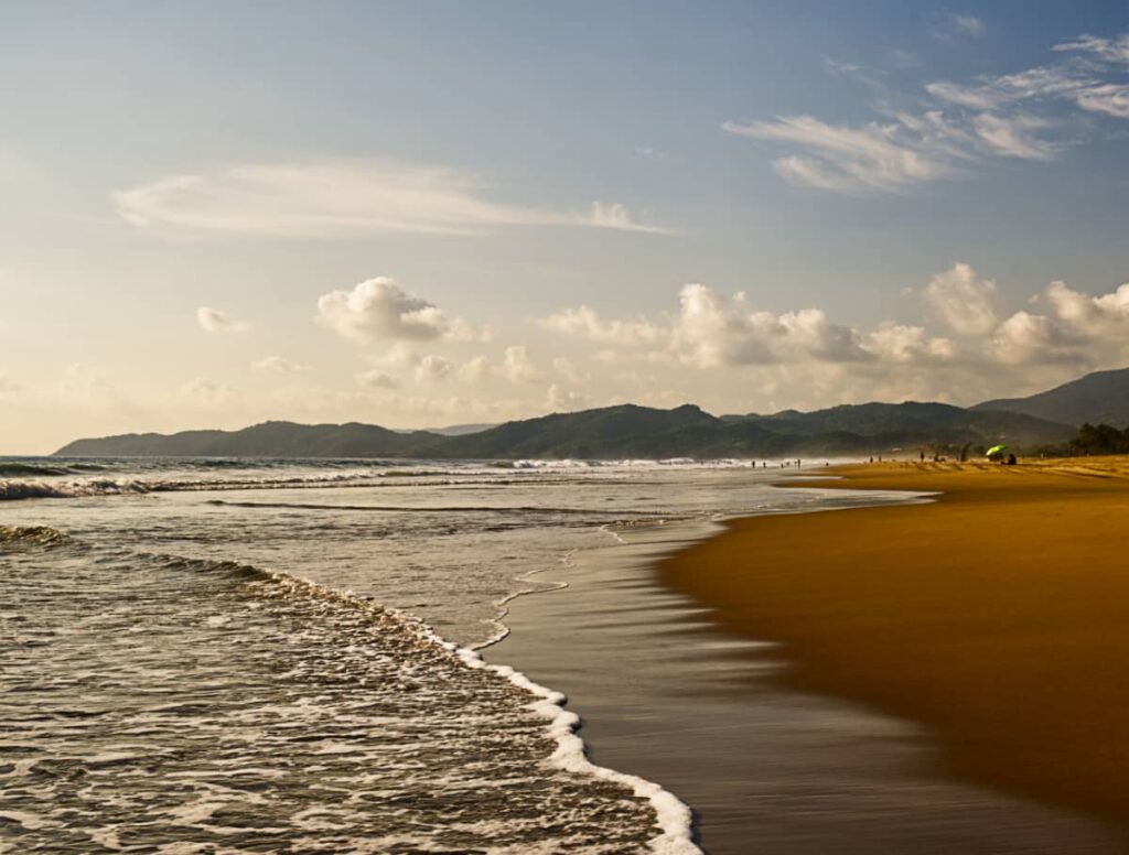 Frothy surf comes on shore to the golden sand beach at Playa Blanca with mountain views and puffy clouds in the background.