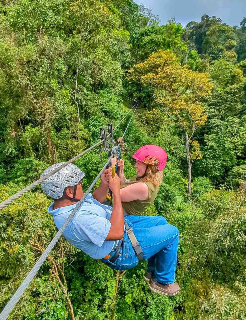 A man and a woman fly along the trees on a zipline in Mindo Ecuador. They are wearing harnesses, helmets, and holding the harness ropes.