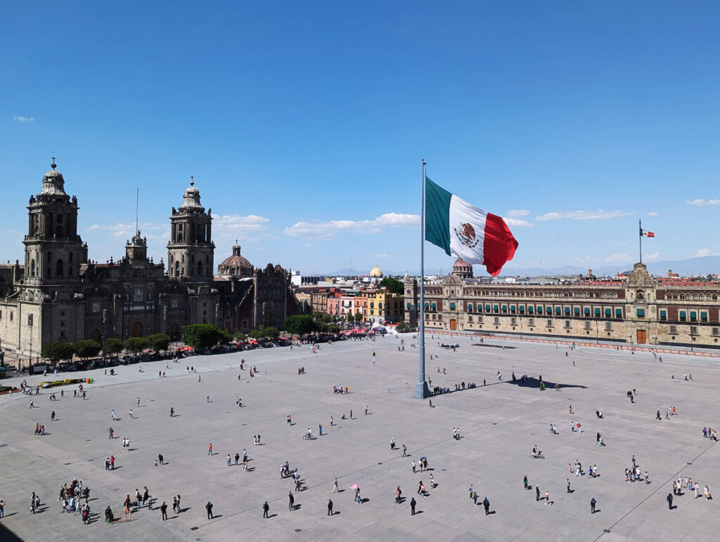 A view from a rooftop in the Zocalo, Mexico City looking at the Cathedral on the left and National Palace on the left. In the center a large Mexican flag flies on a pole while people walk in the Zocalo below.